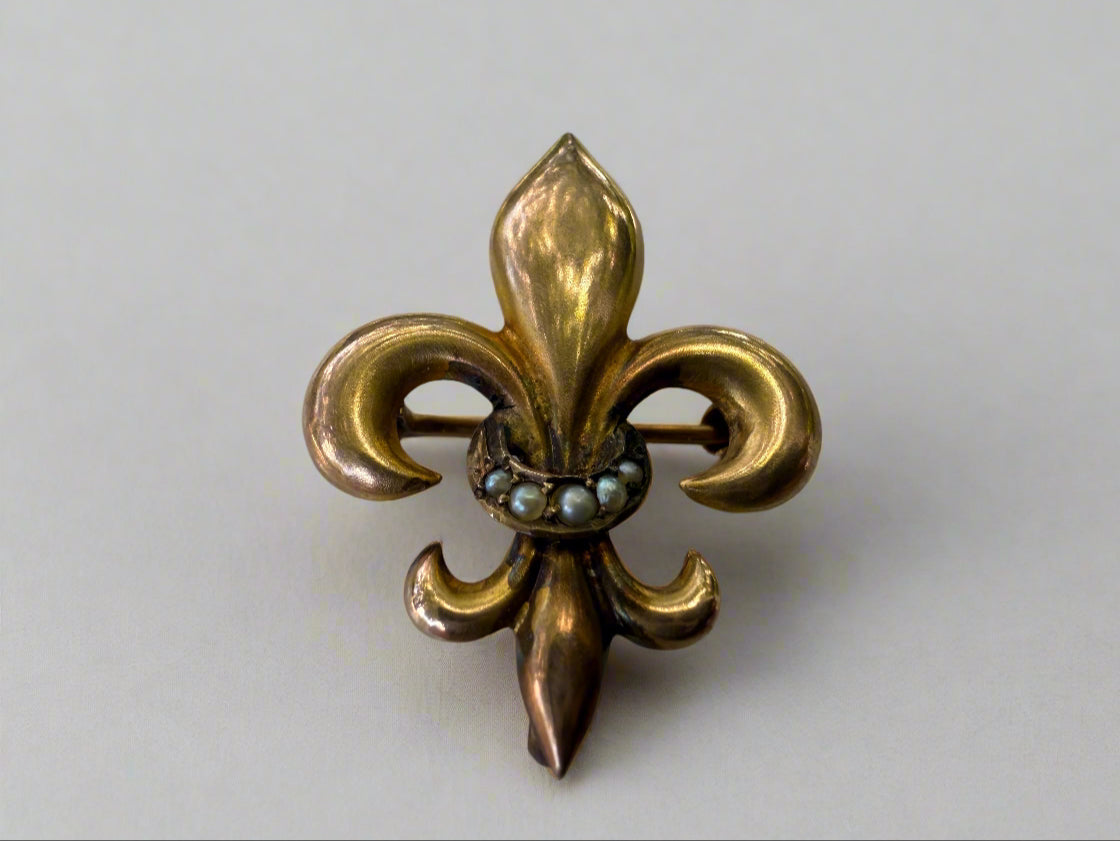 Gold fleur-de-lis brooch with central seed pearls on a light gray background