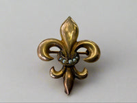 Gold fleur-de-lis brooch with central seed pearls on a light gray background