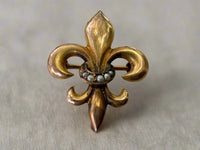 Gold fleur-de-lis brooch with pearl center on a light gray background